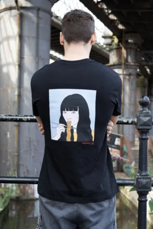 Kanada-Ya | Ramen Near Me | A man with short brown hair, seen from behind, wears a Noodle girl Ichi black T-shirt featuring an illustration of a person eating noodles. Hes standing by a railing under an urban bridge. | London