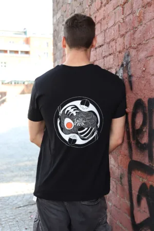 Kanada-Ya | Ramen Near Me | A person with short brown hair stands facing a brick wall, wearing the Onsen Tee Dark. The tee features an abstract circular design on the back with intricate white lines and a central orange dot. This outdoor scene shows buildings in the background. | London