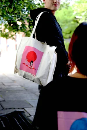 Kanada-Ya | Ramen Near Me | A person outdoors carries the Full Sun Bag, a beige tote with pink artwork of a girl in front of a large red circle. Another individual is partially visible in the foreground amidst lush green trees and a paved path. | London